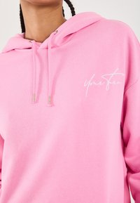 Person, der einen rosa Hoodie mit Kordeln trägt, auf dem in geschwungener weißer Schrift "Dein Zug" auf der Brust steht. Es wird der Oberkörper und der Hals gezeigt.