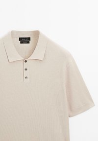 Beige ribstrikket poloshirt lavet af 100 % bomuld, med krave og tre mørke knapper langs knappelisten. Glat tekstur.