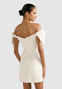 Bijela haljina s otpalim ramenima od strukturiranog satenskog materijala, sa mašnama na rukavima i skrivenim zatvaračem na leđima.