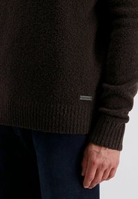 Donkerbruine textuurtrui met ribbels aan de manchetten en onderrand. Heeft een klein metalen label aan de onderzijde. De stof lijkt zacht en zwaar.