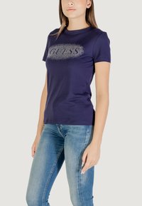 T-shirt in cotone blu navy a maniche corte con un logo "GUESS" scintillante in un design arrotondato sul petto, abbinato a jeans in denim chiaro.