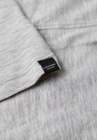 Nærbillede af lysegråt strikket stof med et lille sort mærke, hvor der står "Superdry Premium", syet tæt på kanten.