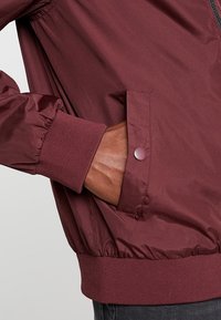 Burgundowa kurtka bomber z elastycznymi mankietami i dołem, z boczną kieszenią zamykaną na napę i gładką, błyszczącą teksturą materiału.