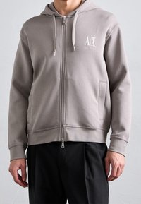 Felpa con cappuccio grigia con cerniera, cappuccio con coulisse, tasche laterali e un piccolo logo bianco. Realizzata in tessuto morbido e strutturato con polsini a coste.