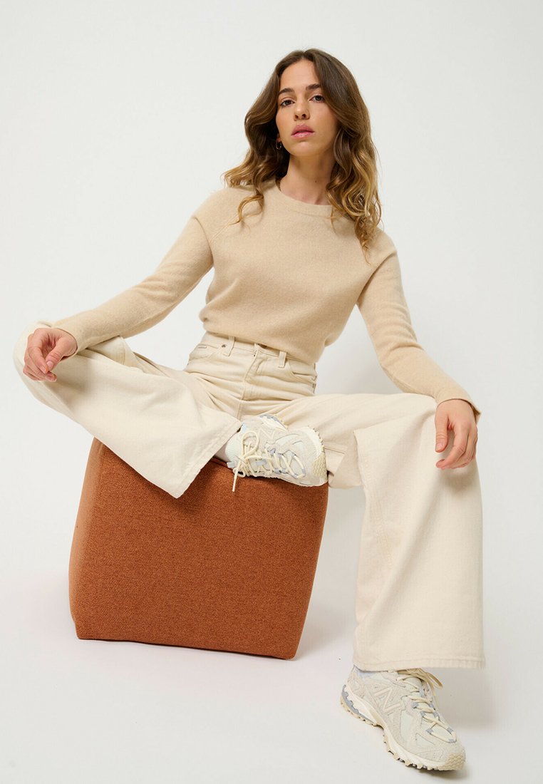 Un pull en tricot beige, un pantalon large crème et des baskets claires portés par une personne assise sur un pouf en mousse texturée de forme cubique marron.