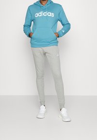 Turkos hoodie med vit "adidas"-logotyp, tillsammans med grå joggers med vita sidosträck. Modellen bär vita sneakers.