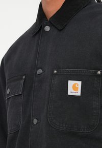Čierna zamatová košeľa s gombíkovým zapínaním, dvoma náprsními vreckami, kovovými cvočkami a bielou štvrtcovou nálepkou s oranžovým logom.