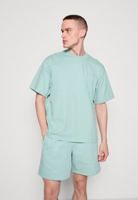Tricou scurt verde mentă, cu mâneci scurte și guler rotund, împreună cu pantaloni scurți asortați. Material din bumbac moale, croială confortabilă, cu un detaliu discret al logo-ului pe piept.