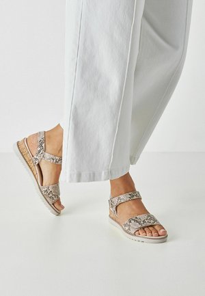 Bruin en beige sandalen met slangenprint, brede bandjes, verstelbare gesp en een gestructureerde zool, gecombineerd met lichtgekleurde broek met wijd uitlopende pijpen.