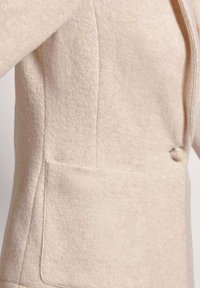 Beige wollen blazer met een gemêleerde structuur, een knoopsluiting met één knop en een grote opgestikte zak aan de linkerkant.