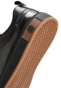 Zapato negro de tipo slip-on con parte superior de cuero liso, suela de goma con textura de espiga y un panel en marrón que muestra un logo.