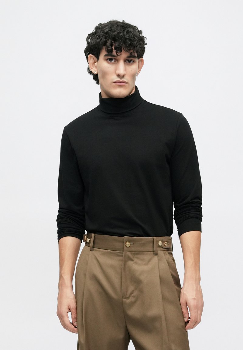 Sort rullekrave trøje med lange ærmer, glat tekstur, parret med høj talje brune bukser med sidelukninger og læg.