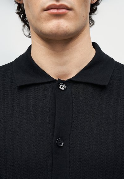 Κοντινό πλάνο στο κάτω μέρος του προσώπου και τον λαιμό ενός ατόμου που φοράει μαύρο υφασμάτινο πουκάμισο με γιακά και κουμπιά.