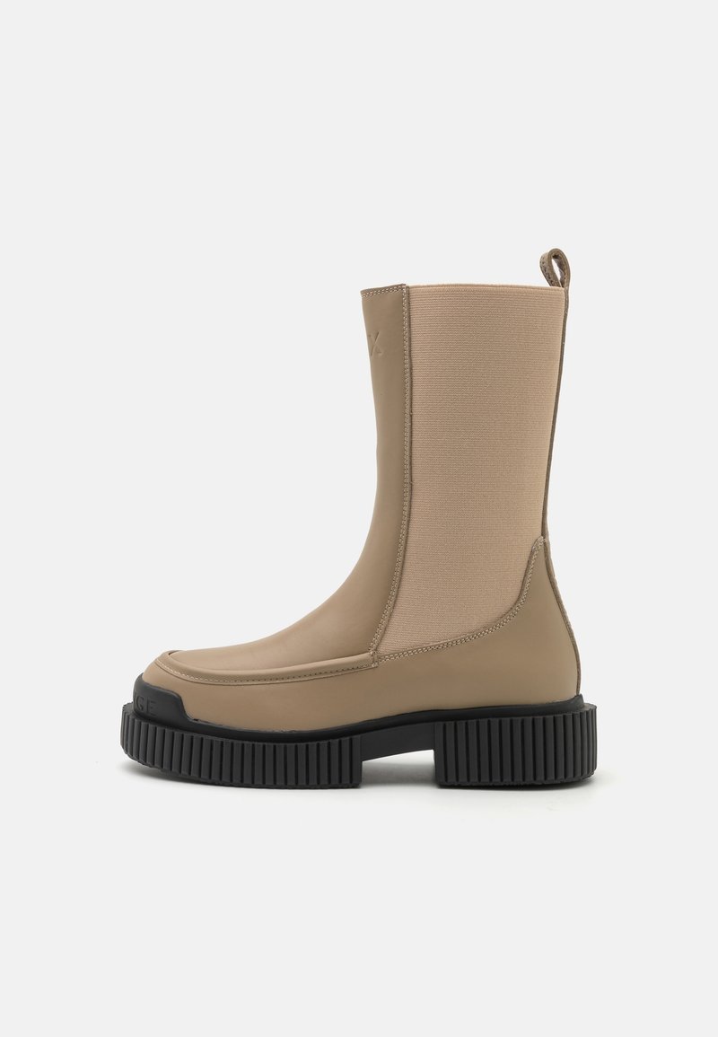 Stivale alla caviglia beige realizzato in un materiale simile alla gomma, con pannelli laterali elastici e una suola piatta nera texturizzata. Include un linguetta per una facile vestibilità.