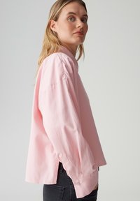 Light pink long-sleeve shirt with a relaxed fit, featuring buttoned cuffs and a high-low hem, paired with dark jeans.