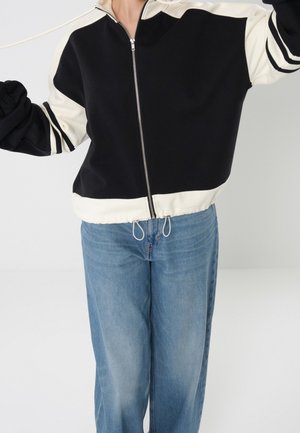 Person trägt eine schwarz-weiß gestreifte Jacke mit Reißverschluss und Kordelzug sowie hellblaue Jeans, steht vor einem schlichten hellen Hintergrund.