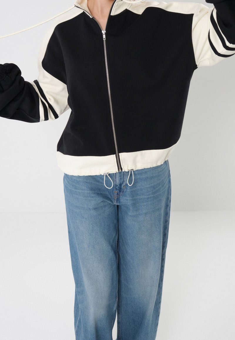 Osoba ubrana w czarno-białą kurtkę na zamek z troczkami oraz jasnoniebieskie jeansy, stojąca na jednolitym jasnym tle.