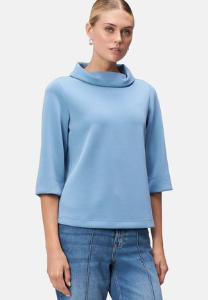 Cowl-Neck-Oberteil aus hellblauem Stoff, mit dreiviertel Ärmeln und lockerer Passform. Glatte Textur und saubere Nähte. Kombiniert mit Jeans.