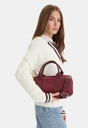 Donna con maglione bianco e jeans chiari tiene una borsa color marrone scuro con manici bordati di nero su sfondo bianco.