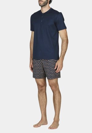 Uomo in piedi a piedi nudi che indossa una maglietta henley blu navy a maniche corte e pantaloncini blu navy con pois beige su uno sfondo uniforme.