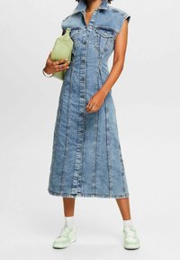 Jeansmaxikjol med knäppning fram, ärmlös design, ljusblå färg och två bröstfickor. Kombinerad med vita och gröna sneakers.
