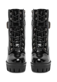 Dvojice černých lesklých bojových bot s tlustými lugovými podrážkami, šněrováním v přední části a přezkami kolem kotníků, z pohledu zepředu.