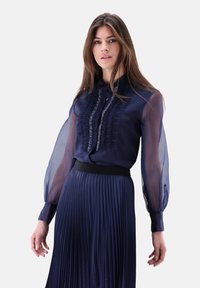 Navy blouse with sheer long sleeves, ruffled front and embellished neckline, paired with a pleated navy skirt and black waistband.