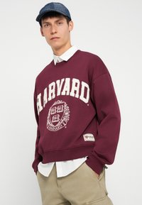 Bordó pulóver nagy "HARVARD" felirattal és emblémával. Pamut anyag, bordás mandzsettákkal és aljjal. Fehér galléros ing felett viselve.