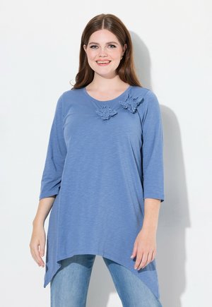 Top de túnica azul con escote redondo, mangas de tres cuartos y detalles decorativos con volantes en el hombro izquierdo. Tela suave y ligera.