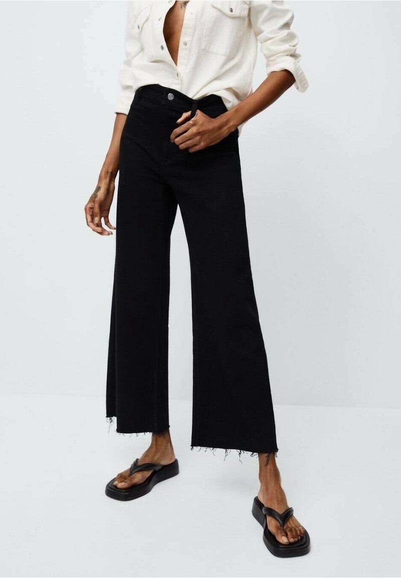 Jeans de pierna ancha negros con dobladillo sin acabar, material de algodón, cintura alta y bolsillos frontales. Combinados con chanclas negras y una camisa blanca de botones.