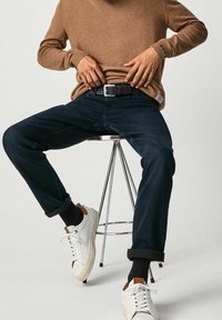 Personne assise sur un tabouret en métal portant un pull marron, un jean bleu foncé avec les ourlets retroussés, des chaussettes noires et des baskets blanches avec des accents marron.