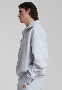 Grå hettejakke med glidelås og høy krage, elastiske mansjetter og en brodert logo i oransje. Myk stoff med en avslappet passform.