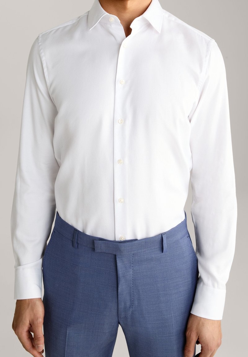Homme portant une chemise blanche ajustée rentrée dans un pantalon de costume bleu sur mesure, debout devant un fond neutre.