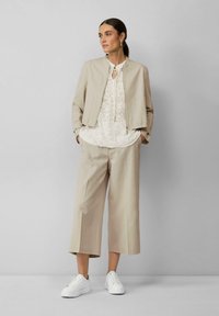 Beige cropped jasje met een gladde textuur, een witte bloemen geborduurde top en brede beige broeken, gecombineerd met witte sneakers.
