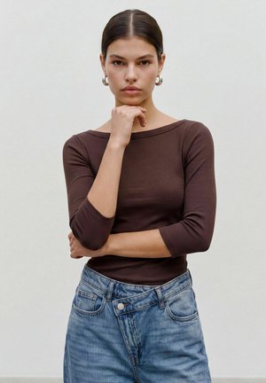 Jeune femme aux cheveux foncés vêtue d'un haut marron à manches longues et d'un jean bleu, se tient debout avec une main sous le menton et l’autre croisée sur la taille.