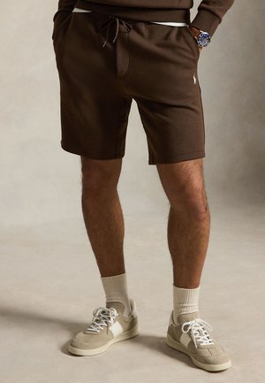 Underkroppen af en person iført brune shorts med snøre, beige sneakers med hvide snørebånd, lyse sokker og et armbåndsur med blå urskive.