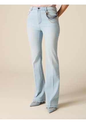 Jeans azzurri a zampa con catene decorative in oro e tessuto sulla tasca, indossati con décolleté a punta, in piedi contro uno sfondo beige.