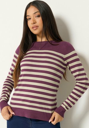 Giovane donna con maglione a righe viola e beige a maniche lunghe, in piedi contro un muro bianco mentre sistema l'orlo inferiore con le mani.