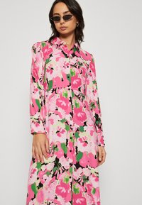 Long button-up dress made of lightweight fabric. Features a vibrant floral pattern in pink, green, and black. Long sleeves, collar design.
