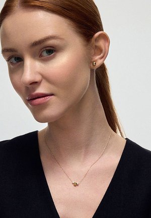 Złoty naszyjnik z małym sferycznym wisiorkiem, noszony przez modelkę o jasnej skórze i rudych włosach, zestawiony z dopasowanymi małymi złotymi kolczykami.