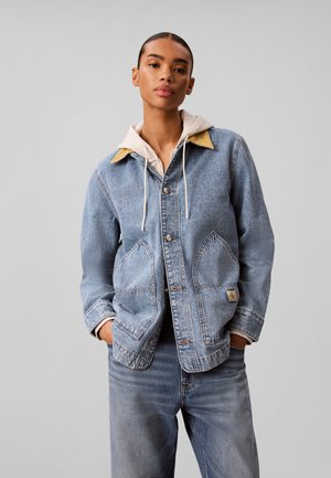 Junge Person trägt eine hellblaue Jeansjacke mit großen Taschen über einem beigen Hoodie und passenden Jeans, steht mit den Händen in den Taschen.