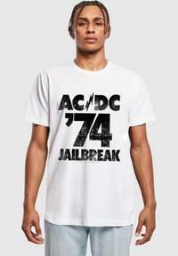 Hvid bomulds t-shirt med sort distressed tekst: "AC/DC '74 JAILBREAK." Korte ærmer, standard pasform. Minimalistisk design.
