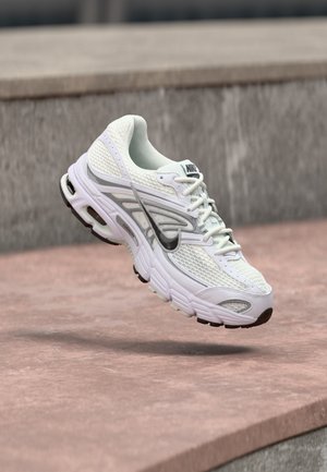 Hvit og grå Nike joggesko svevende i luften over rødbrun asfalt med uskarpe betongtrinn i bakgrunnen.