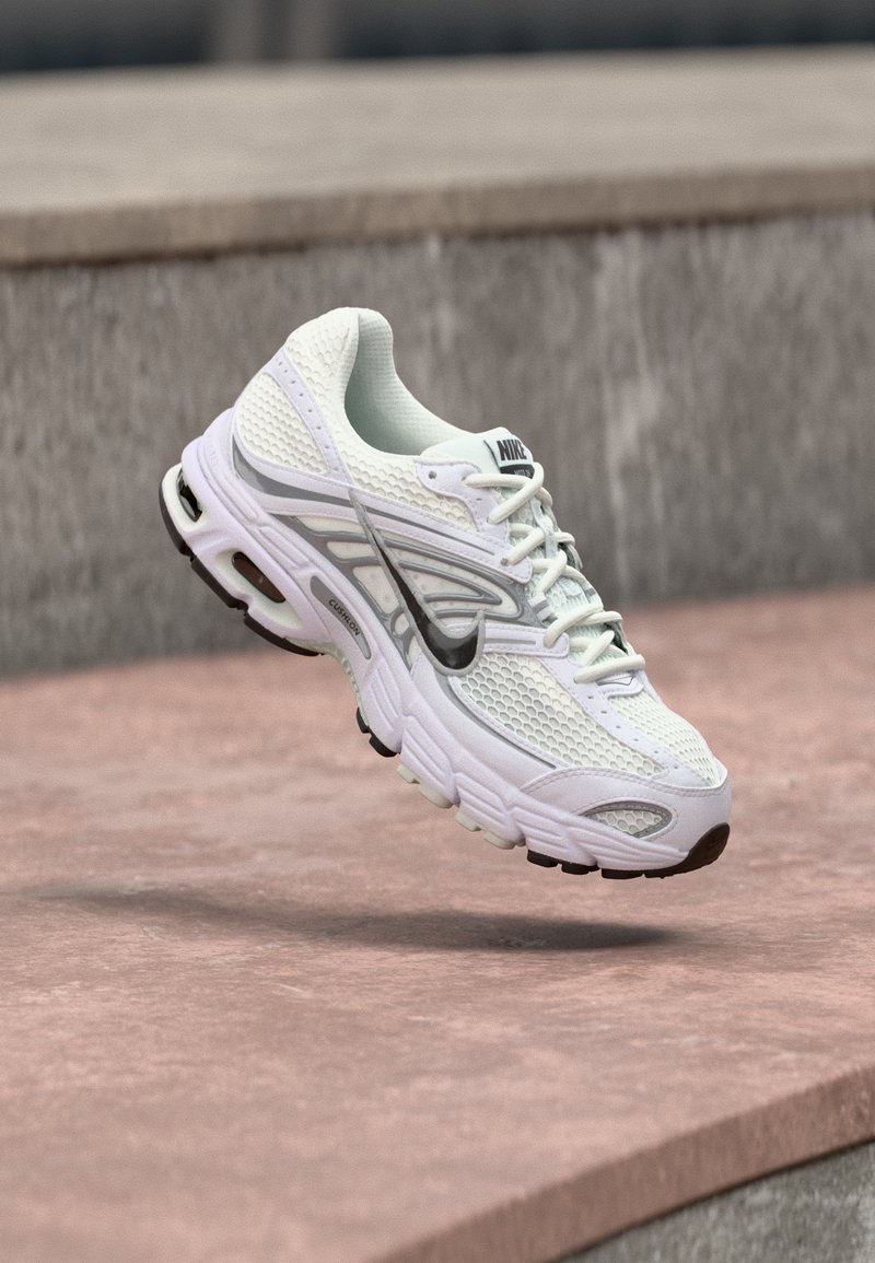 Sapatilha de corrida Nike branca e cinza suspensa no ar acima de um pavimento castanho avermelhado, com degraus em concreto desfocados ao fundo.