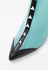 Escarpin en cuir verni turquoise avec un bout pointu ; il présente une bordure noire et une rangée décorative de clous en pyramide argentés sur le côté.