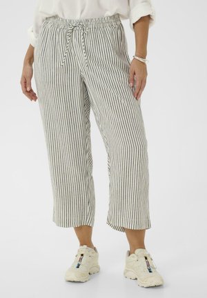 Kvinde iført korte, hvide bukser med sorte lodrette striber, snøre i taljen, hvide sneakers og en hvid top med opkørte ærmer.