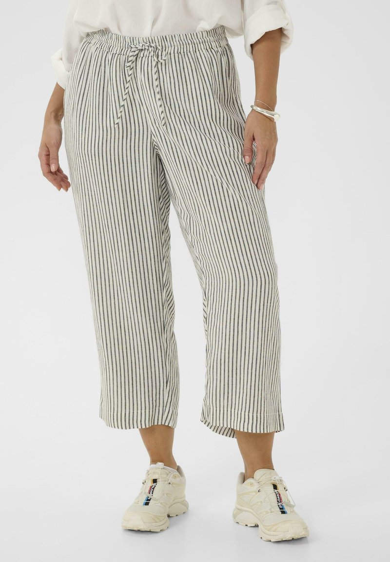 Nő, aki fehér, fekete függőleges csíkos, rövidített nadrágot, húzózsinóros derekúdarabot, fehér sportcipőt és felhajtott ujjú fehér felsőt visel.