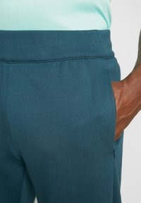 Närbild av en person som bär turkosa sweatpants med en hand placerad i den dragkedjade sidofickan.