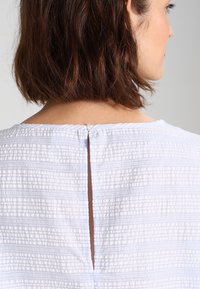 Weißes, strukturiertes Blusenshirt mit rundem Halsausschnitt und kleiner Knopfverschluss auf der Rückseite. Der Stoff zeigt ein dezentes Streifenmuster.