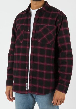 Man draagt een zwart-rood geruit overhemd met open knopen over een wit T-shirt en lichtblauwe spijkerbroek, staand voor een witte achtergrond.